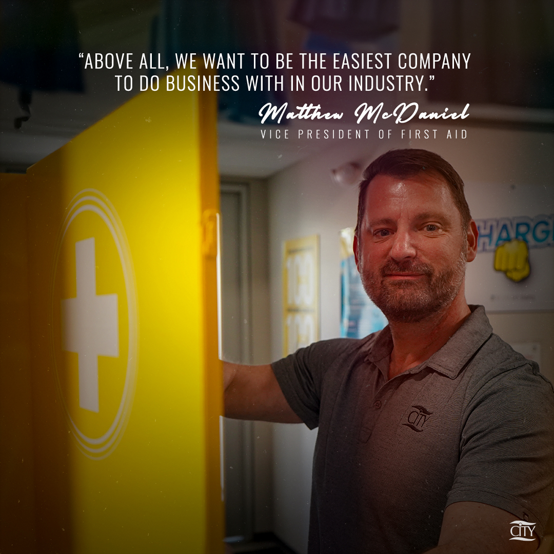 Matthew McDaniel, Vice President of First Aid at CITY, stands beside an open yellow first aid cabinet with a white cross symbol, smiling slightly. A quote above him reads, “Above all, we want to be the easiest company to do business with in our industry.”
